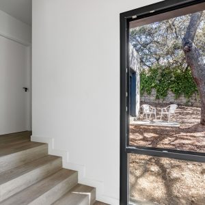 Vista interior de una escalera con peldaños de madera clara y un gran ventanal de aluminio negro que enmarca un jardín natural de encinas en Madrid, diseñada por Espacio Acorde.