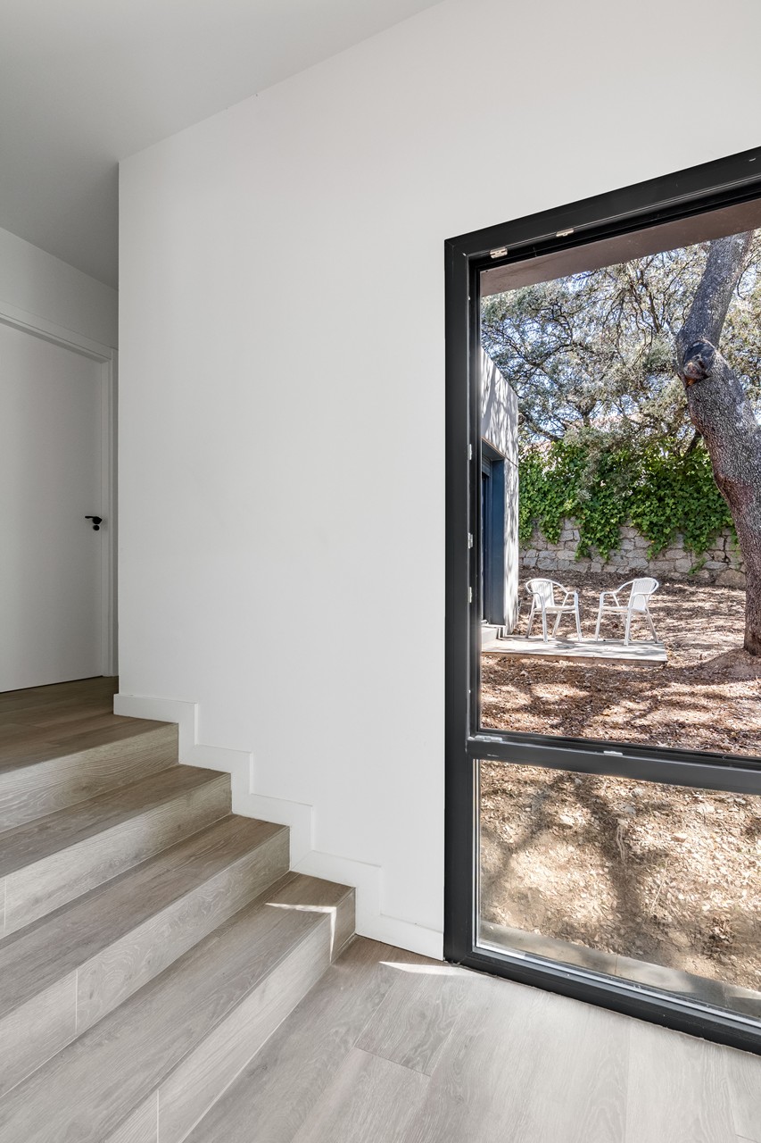 Vista interior de una escalera con peldaños de madera clara y un gran ventanal de aluminio negro que enmarca un jardín natural de encinas en Madrid, diseñada por Espacio Acorde.