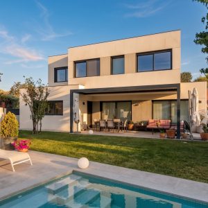Fachada posterior de vivienda unifamiliar de dos plantas en Boadilla del Monte, con porche cubierto, zona de comedor exterior, jardín con césped y piscina integrada, proyectada por Espacio Acorde.