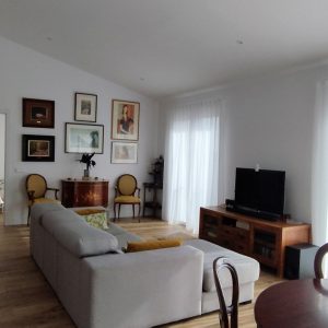 Vista interior del salón de una vivienda unifamiliar en El Escorial (Madrid), con sofá gris, mueble de televisión en madera, pared con cuadros, estantería de obra blanca y paso abierto hacia dormitorio infantil, por Espacio Acorde.
