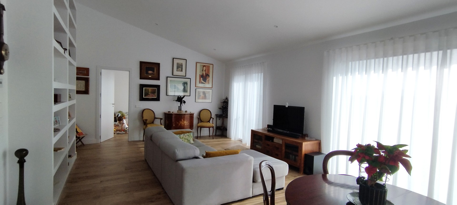 Vista interior del salón de una vivienda unifamiliar en El Escorial (Madrid), con sofá gris, mueble de televisión en madera, pared con cuadros, estantería de obra blanca y paso abierto hacia dormitorio infantil, por Espacio Acorde.