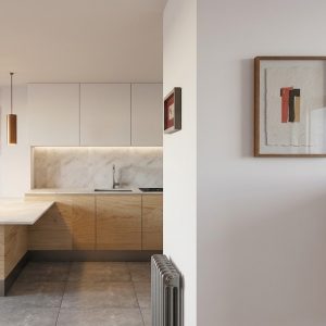 Vista de transición desde el recibidor con aparador de madera labrada y cuadro enmarcado hacia la cocina con muebles de madera y mármol, luminarias cilíndricas y comedor al fondo en piso de Alcalá Norte (Madrid), por Espacio Acorde.