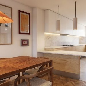 Vista diagonal de la zona de comedor con mesa de madera y lámpara cónica, junto a cocina semiabierta en L con península de mármol, armarios de madera y ventana exterior en piso de Alcalá Norte (Madrid), por Espacio Acorde.