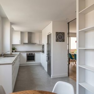 Fotografía de cocina reformada en piso de Madrid, con mobiliario blanco en L, puerta corredera acristalada hacia el salón, estantería de madera a medida y ventana a la terraza, por Espacio Acorde.
