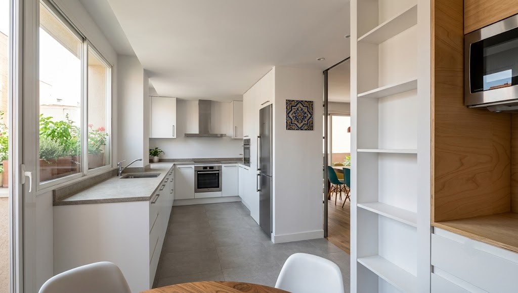 Fotografía de cocina reformada en piso de Madrid, con mobiliario blanco en L, puerta corredera acristalada hacia el salón, estantería de madera a medida y ventana a la terraza, por Espacio Acorde.